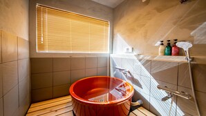Un baño moderno con suelo de madera, una bañera roja, una ventana con persianas y una zona de ducha con cabezal de ducha manual.