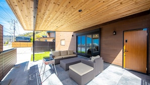 Un patio moderno al aire libre con techo de madera, un área de estar con sofá y reposapiés, y una puerta a la derecha.