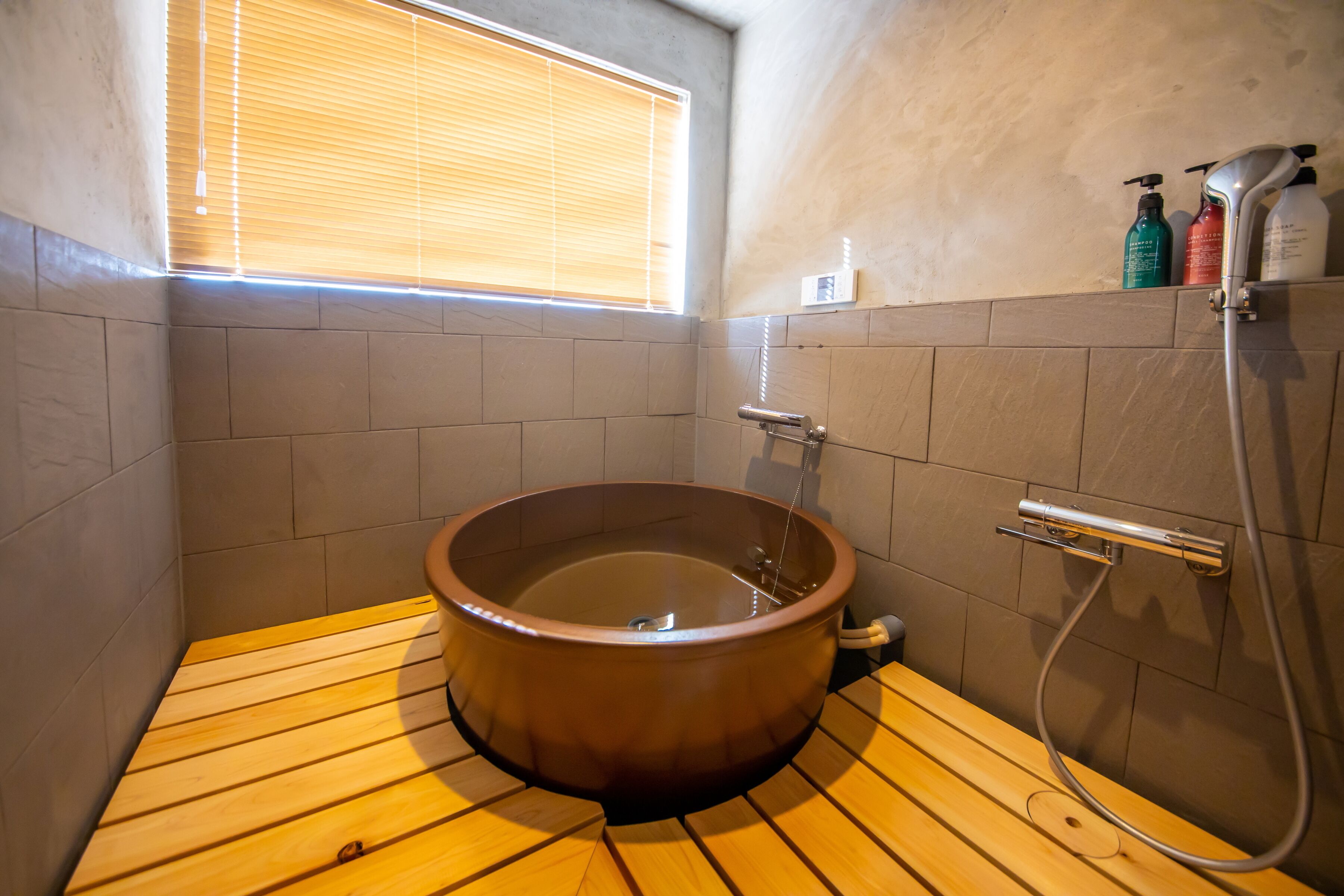 A wooden sauna with a tiled floor, a window with blinds, and a shower area with bottles of shampoo and conditioner.