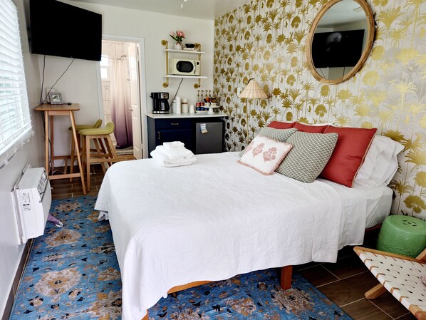 A neatly arranged bedroom with a bed, a desk, a chair, and a small kitchenette.