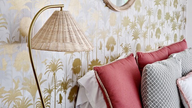 Una stanza con carta da parati decorata, uno specchio rotondo, una lampada da terra e un letto con cuscini.