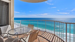 Un balcon avec une table ronde et des chaises, donnant sur une plage aux eaux bleu cristallin et agrémentée d’une balustrade blanche.