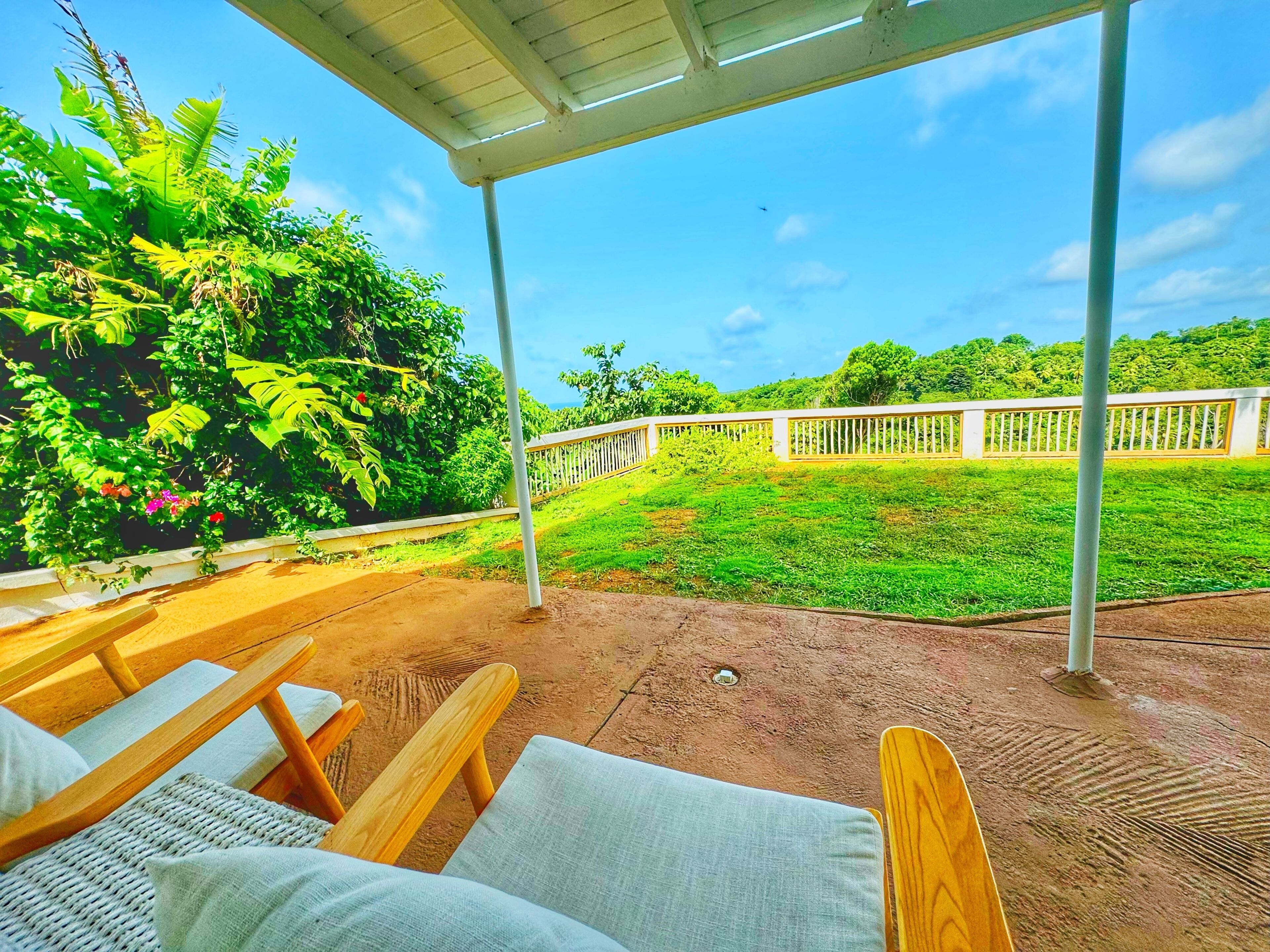 A covered outdoor seating area with wooden chairs and a concrete floor, surrounded by lush greenery and a white fence.