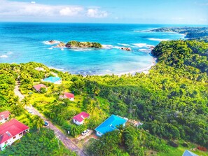 An aerial view of a coastal area with lush greenery, a beach, and several buildings with red roofs. - Blue Whale Villa (Calibishie)