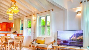 A modern living room with a sofa, dining table, and chairs, a television, and large windows with curtains. - Blue Whale Villa (Calibishie)