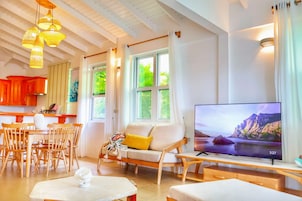 Una sala de estar moderna con sofá, mesa de comedor y sillas, televisor y ventanas grandes con cortinas.
