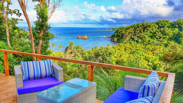 A deck with wicker furniture, a glass table, and a view of the ocean and greenery. - Blue Whale Villa (Calibishie)