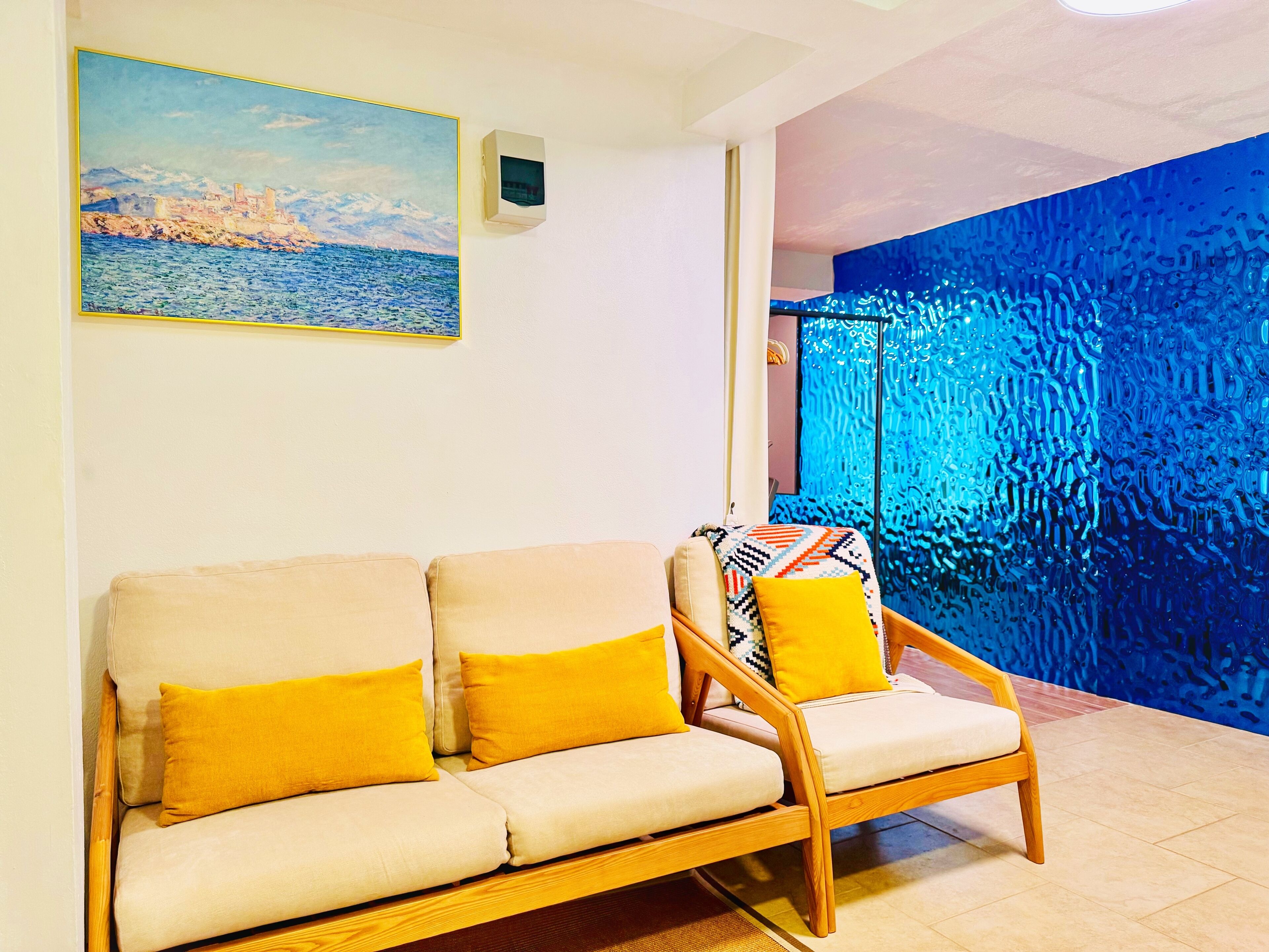 A living room with a beige sofa and a wooden chair with yellow cushions, a painting of a coastal scene, and a blue textured wall.
