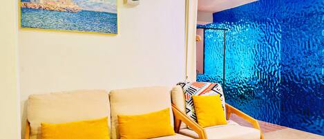 A living room with a beige sofa and a wooden chair with yellow cushions, a painting of a coastal scene, and a blue textured wall.