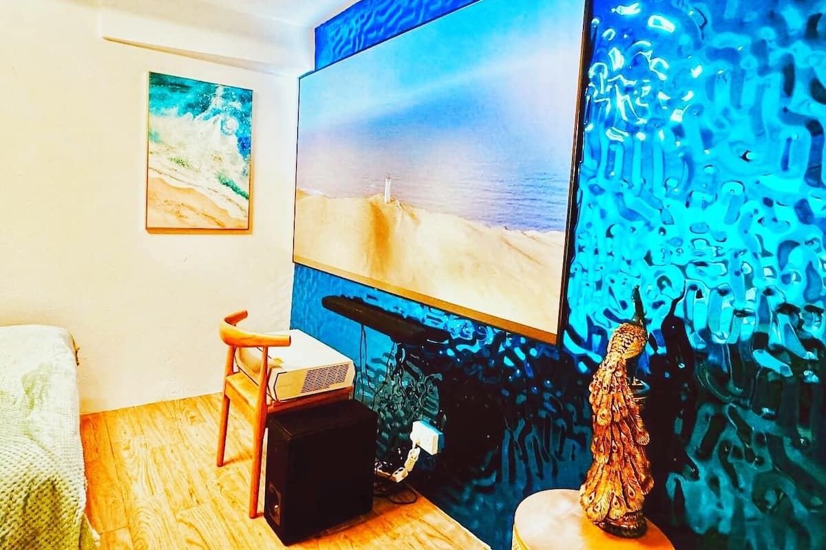A room with a large flat-screen TV mounted on a blue textured wall, a small wooden table with a speaker, a decorative statue, and a painting on the wall.