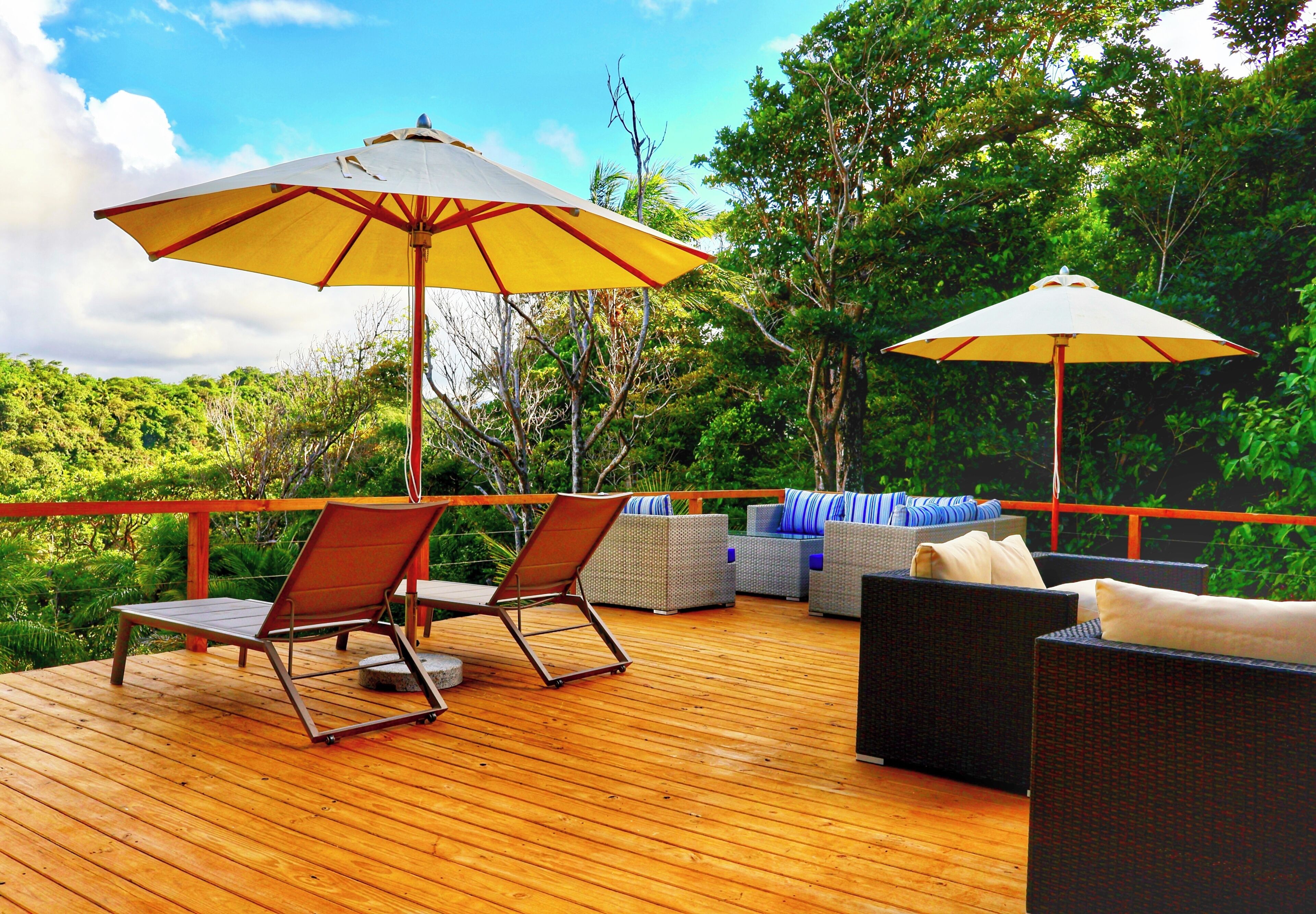 A wooden deck with outdoor furniture, including two lounge chairs and a sofa, and two umbrellas providing shade.