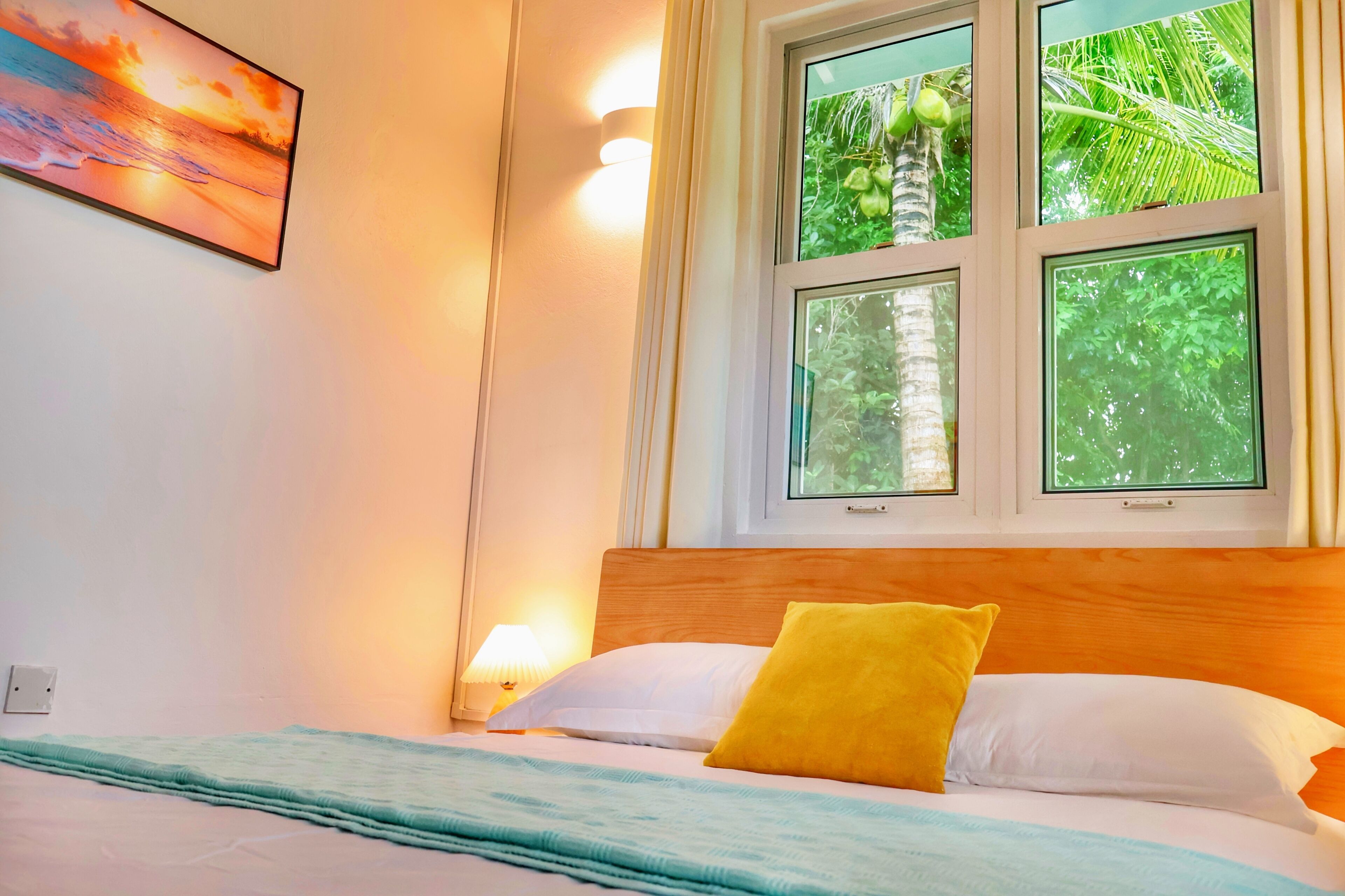 A bedroom with a bed, a window with a view of greenery, a wall-mounted lamp, and a painting.