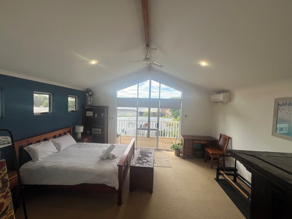 A bedroom with a bed, a desk, a chair, a ceiling fan, and a view of the outside.