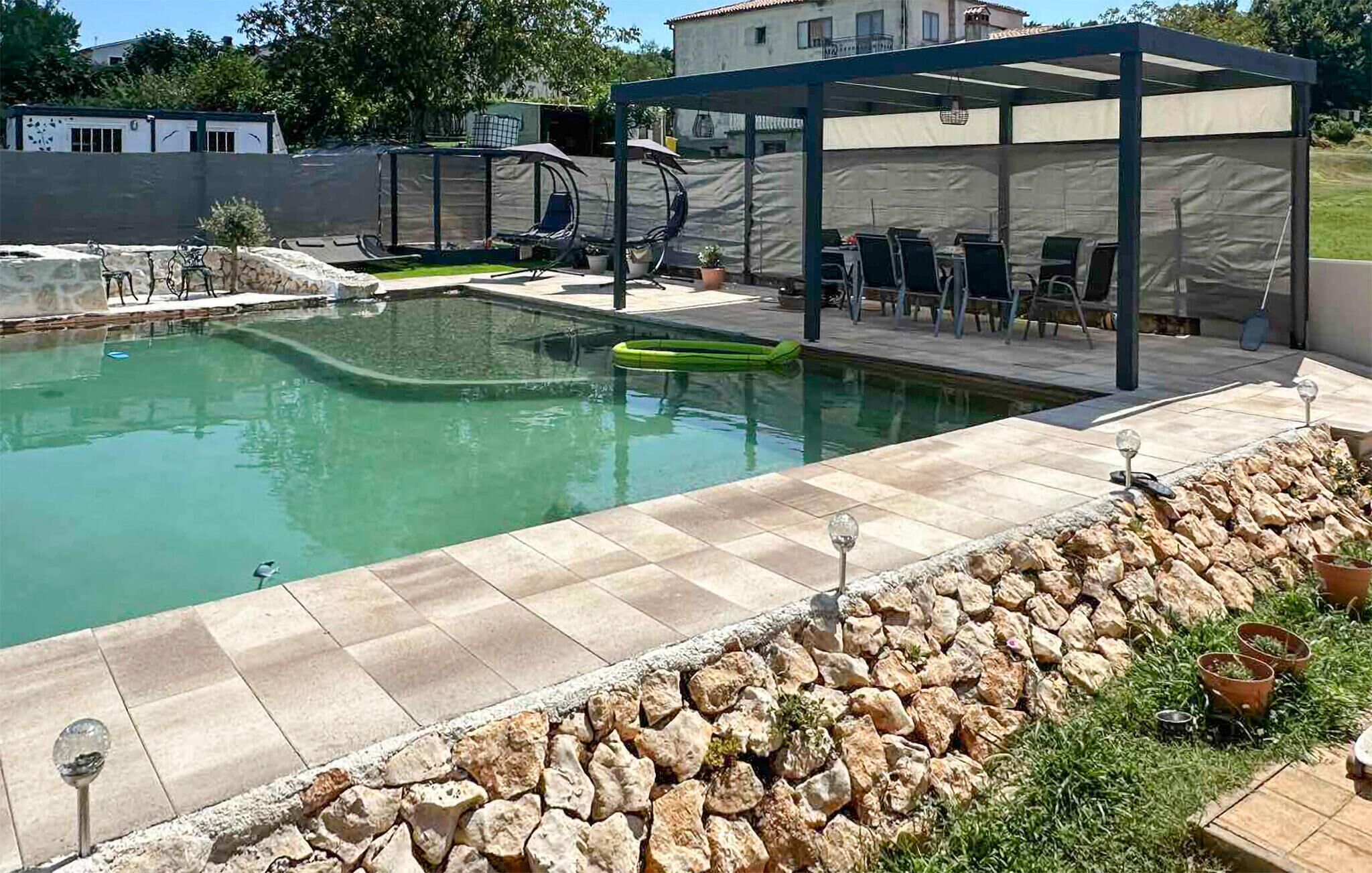 Una piscina con borde de piedra, zona de estar cubierta y un trampolín.