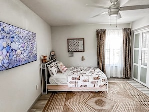 A bedroom with a bed, a ceiling fan, a large abstract painting, a framed chessboard, and a view of the outdoors through a window with curtains.