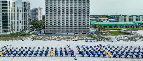 A beachfront hotel with multiple floors, a sandy beach with umbrellas, and a clear view of the ocean.