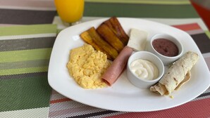 A breakfast plate with scrambled eggs, ham, fried plantains, a burrito, and a glass of orange juice on a striped tablecloth. - Seven Days Hotel B&B (San Pedro Sula)