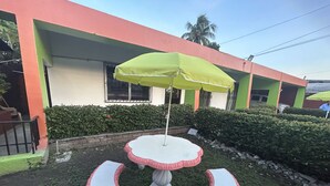 A patio with a table and chairs, a green umbrella, and a building with a red and green facade. - Seven Days Hotel B&B (San Pedro Sula)