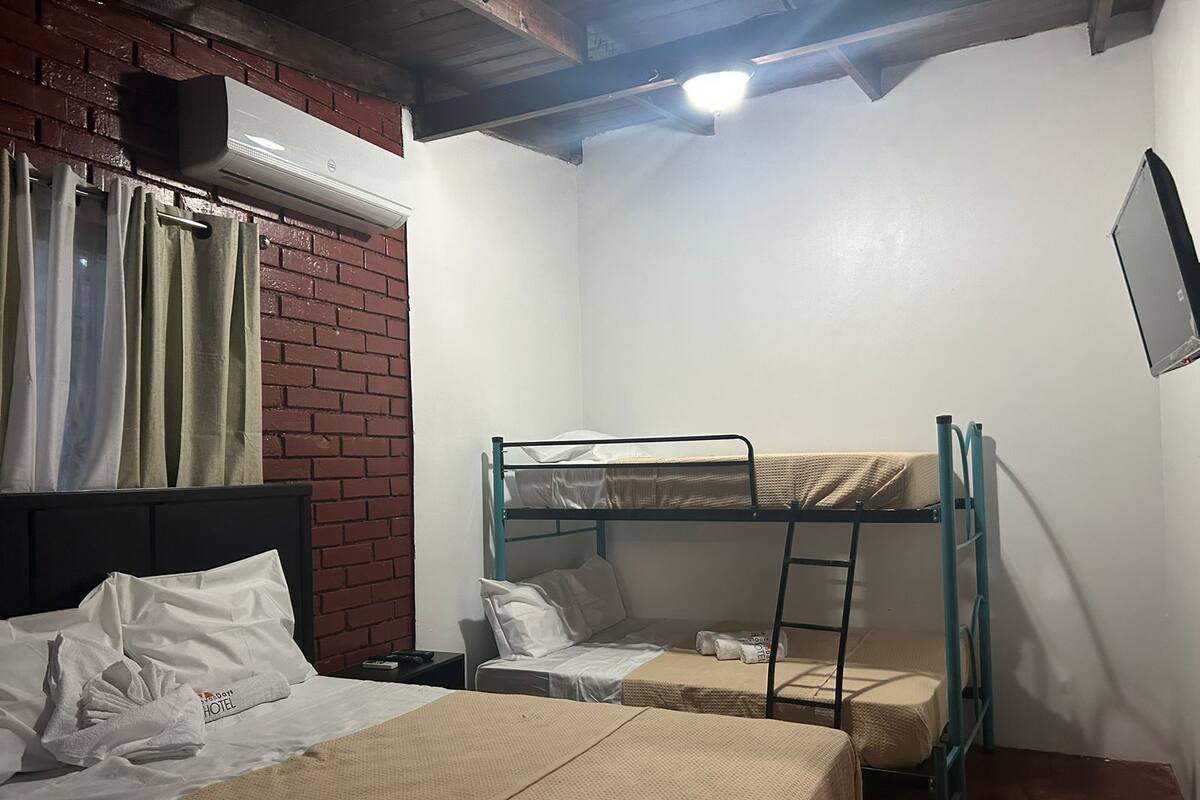 Une petite chambre d’h ôtel, de taille modeste, avec deux lits, un mur en briques, une fenêtre avec des rideaux et un ventilateur de plafond.