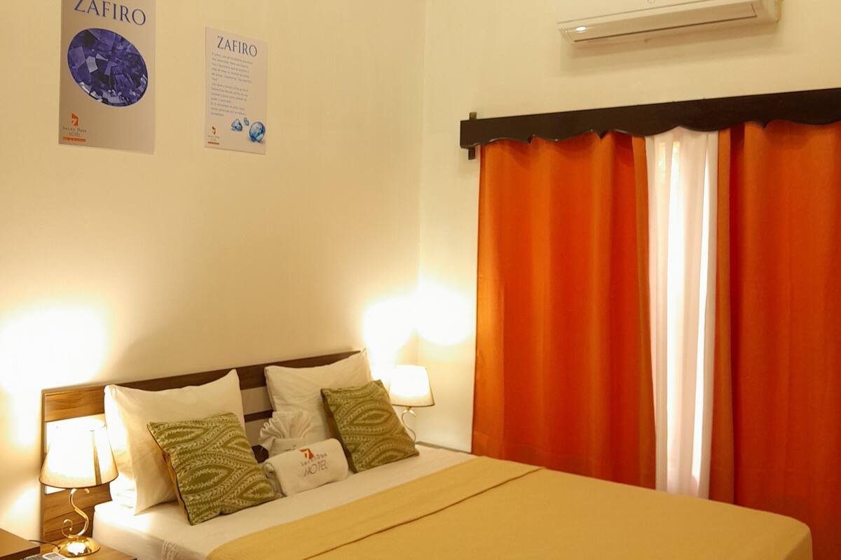 Une chambre d’hôtel comprenant un lit, deux lampes de chevet, une table de nuit, un climatiseur fixé au mur et deux affiches d’information.