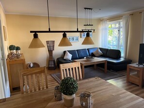 Una sala de estar con un sofá, una mesa de comedor, sillas, un televisor y una ventana con cortinas.