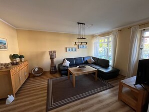 Ein Wohnzimmer mit einer Couch, einem Couchtisch und einem Fernseher. Es gibt ein Holzregal und einen kleinen runden Tisch mit einem Korb.