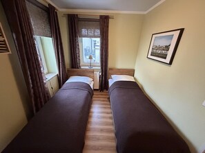 Ein Zimmer mit zwei Einzelbetten, einem fenster mit Vorhängen, einem Heizkörper und einem gerahmten Bild an der Wand.