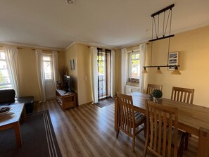 Ein Essbereich mit einem Holztisch und Stühlen, einem Fernseher auf einem Schrank und Blick ins Freie durch ein Fenster.