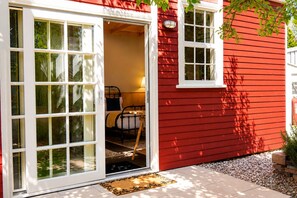 Una casa roja con puerta blanca, una cama con estructura negra y una mesita.