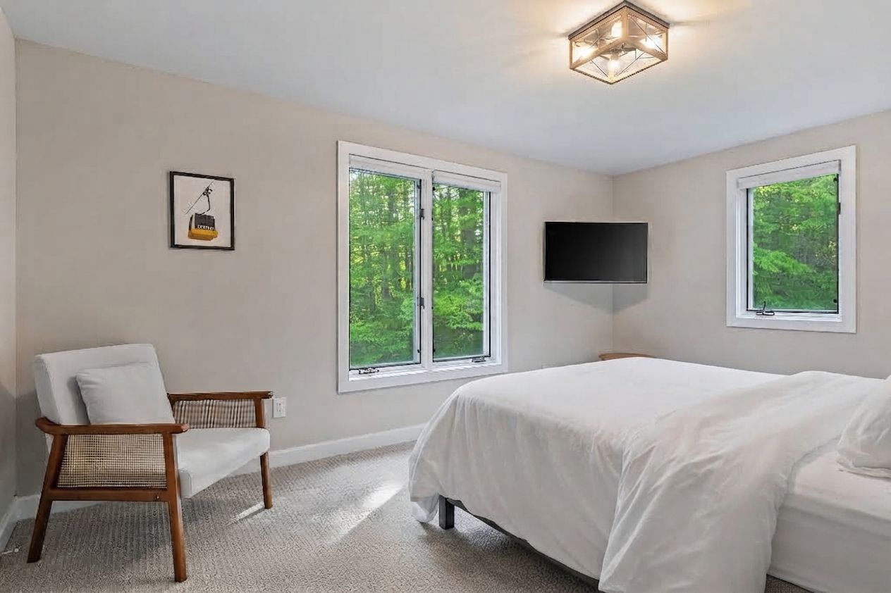 A bedroom with a bed, a chair, a television, and two windows.