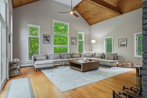 A spacious living room with a large sectional sofa, a coffee table, and multiple windows offering views of greenery. - Hot Tub, Game Room, Pool Table | Weekend Stratton Shuttle, 5BR (Winhall)