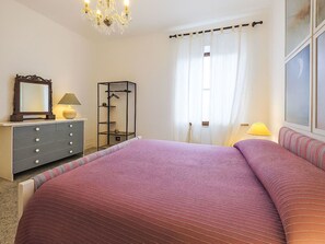 Ein Schlafzimmer mit Bett, Kommode, Spiegel, Lampe und einem Fenster mit Vorhängen.