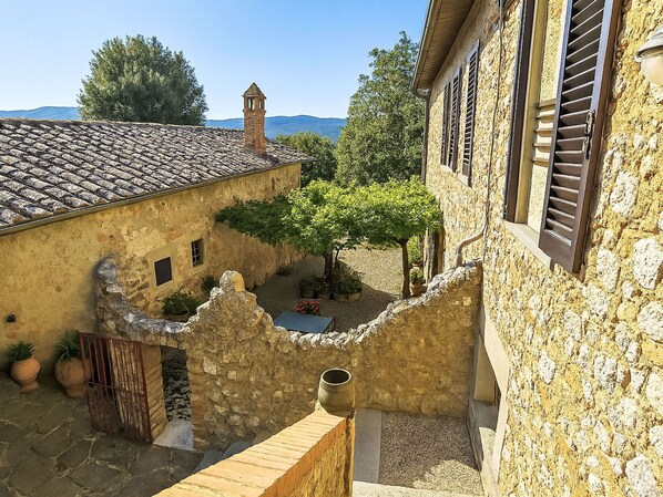 A courtyard with stone buildings, a small tower, and a view of the sea.