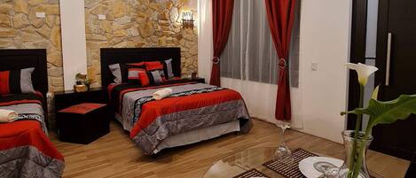 A hotel room with two beds, a stone wall, a chandelier, a dining table with a vase, and a window with red curtains.