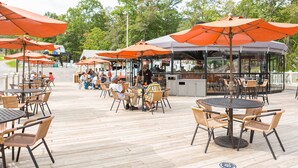 An outdoor patio area with round tables and chairs, orange umbrellas, and a bar area with a view of the trees.