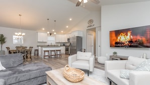 A modern living room with a grey sofa, a white armchair, and a wooden coffee table. There is a kitchen in the background with white cabinets and a dining area.