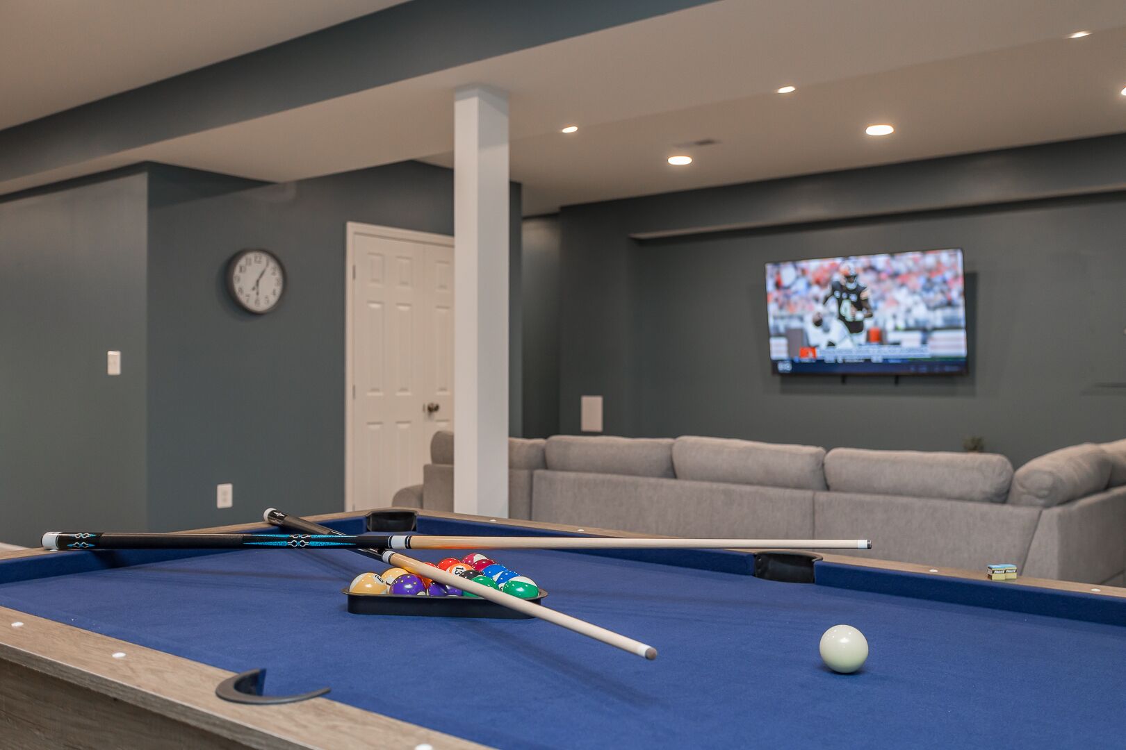 A pool table with balls and cues, a television on the wall, and a gray sofa in a room with gray walls and a white door.