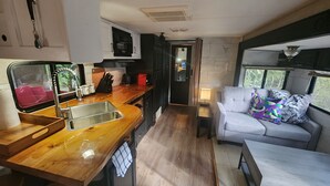 A compact kitchen with a sink, a microwave, and a toaster. There is a wooden countertop and a window with curtains. A white sofa with patterned cushions is visible. - Forest Cabin - Hot Tub - Fast WiFi - Fire Pit On Site (Duncan)