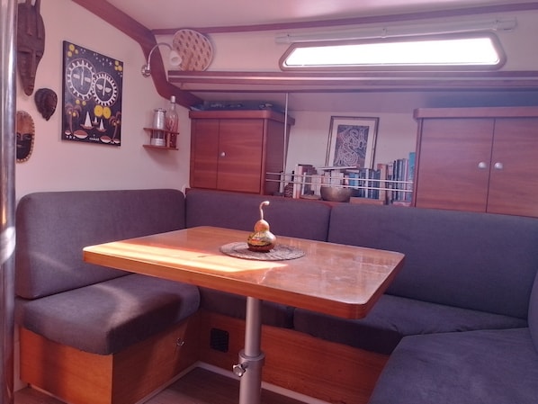 A compact living area with a wooden table, cushioned seating, a small shelf with decorative items, and a wall-mounted light fixture.