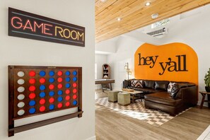 Une pièce avec un panneau néon indiquant « SALLE DE JEUX » et un jeu de société GoSports. On y trouve un grand mur orange sur lequel est écrit « hey y'all », un canapé en cuir marron et un plafond en bois.