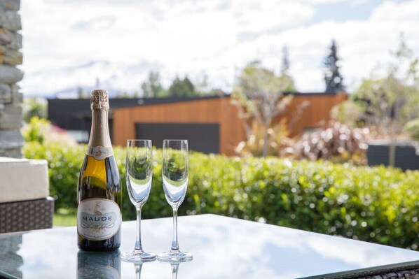Eine Flasche Maude-Wein und zwei Gläser auf einem Tisch, mit Blick auf Grünflächen und Gebäude im Hintergrund.