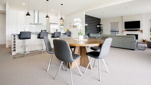 A modern kitchen with white cabinets, a dining area with a wooden table and grey chairs, a grey sofa, and a flat-screen TV mounted on the wall. - Daniels Delight (Wānaka)