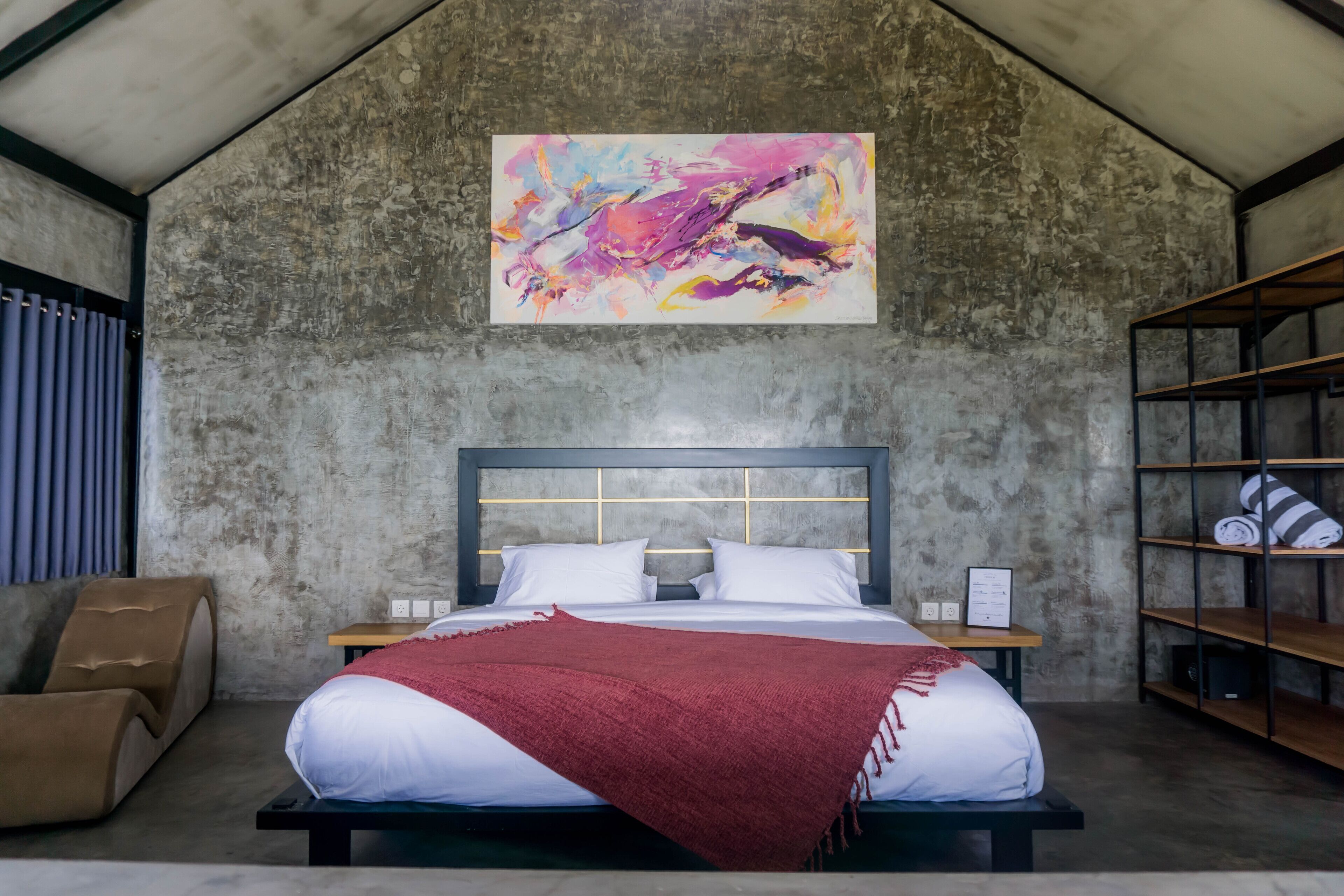 Un dormitorio con una cama grande, un cabecero con estampado de cuadrícula, una manta roja, una mesita de noche de madera, un estante con toallas enrolladas y un cuadro abstracto colorido en la pared.