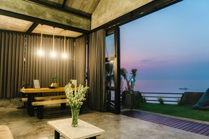 A modern interior with a wooden bench, a table with a vase of flowers, and a view of the sea at dusk. - EJ House (Buleleng)