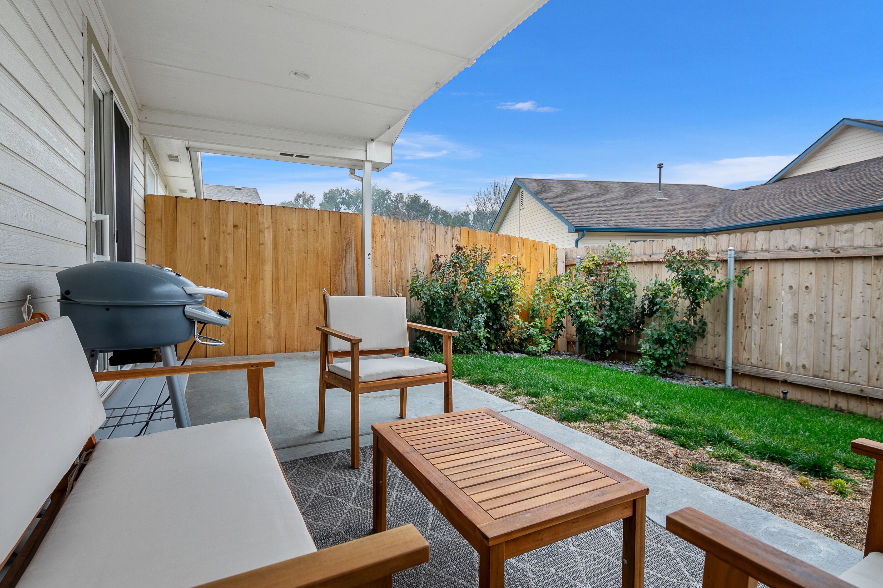Une terrasse dotée de meubles en bois, d’un barbecue et offrant une vue sur une cour arrière avec une clôture en bois et de la verdure.