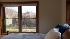 A bedroom with a large window, a bed with white and blue bedding, and a view of trees and hills outside. - Mee Mee's Cabin Retreat- River, nature, small town, Hot Tub
 (Genoa)