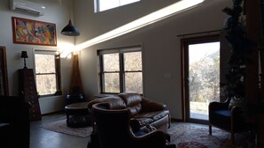 A spacious living room with a leather sofa, a coffee table, and a fireplace. There are large windows allowing natural light and views of the outdoors. - Mee Mee's Cabin Retreat- River, nature, small town, Hot Tub
 (Genoa)