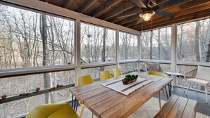 Una mesa de comedor de madera con sillas amarillas, un ventilador de techo y grandes ventanales con vistas a un bosque.