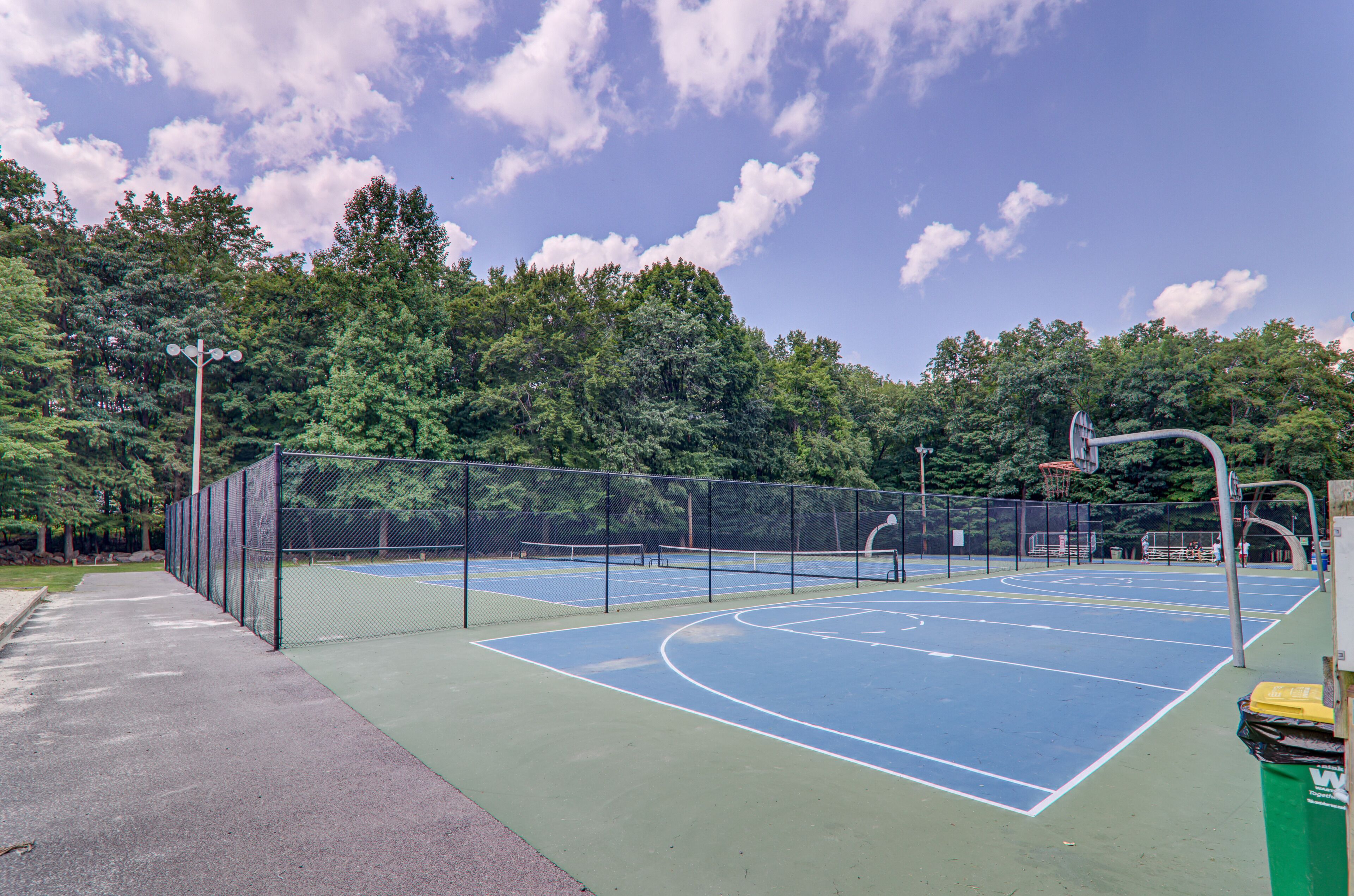 An outdoor basketball court with a fence, basketball hoops, and a trash bin.