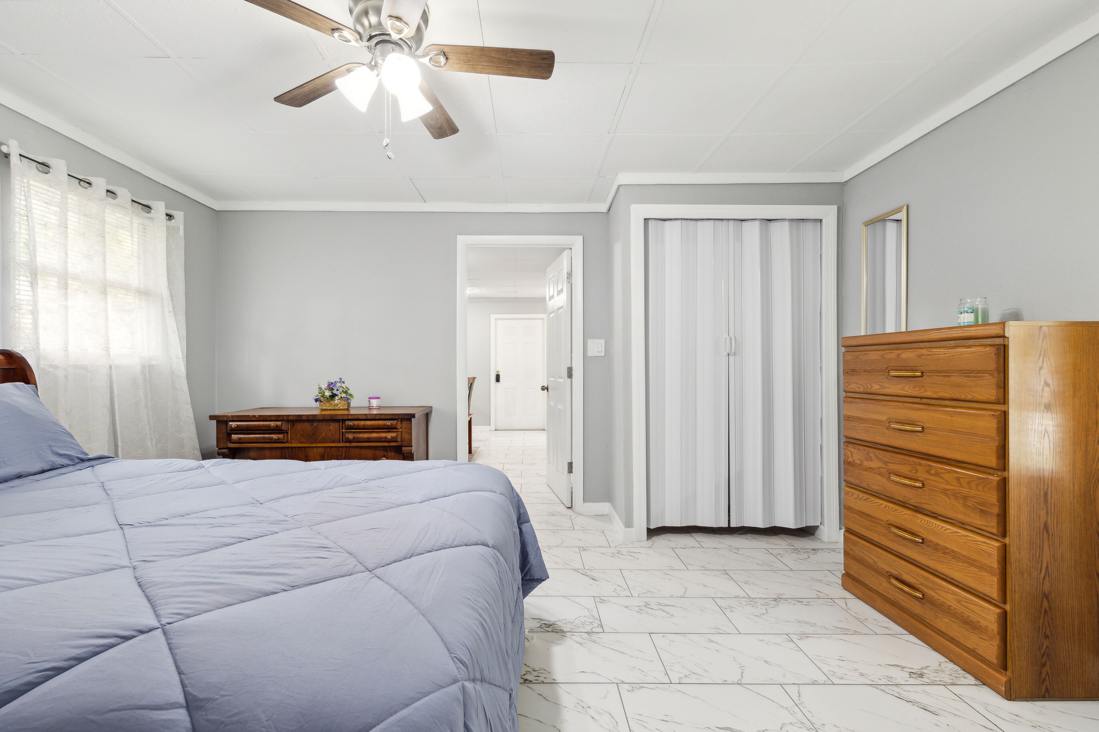 Un dormitorio con cama, cómoda de madera, ventilador de techo y puerta que da a otra habitación.
